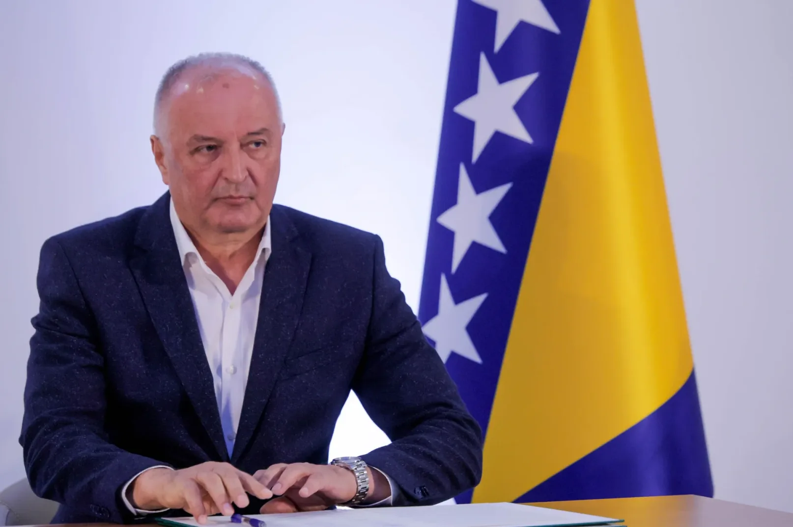 Političar u tamnom odijelu sjedi pored zastave Bosne i Hercegovine.
