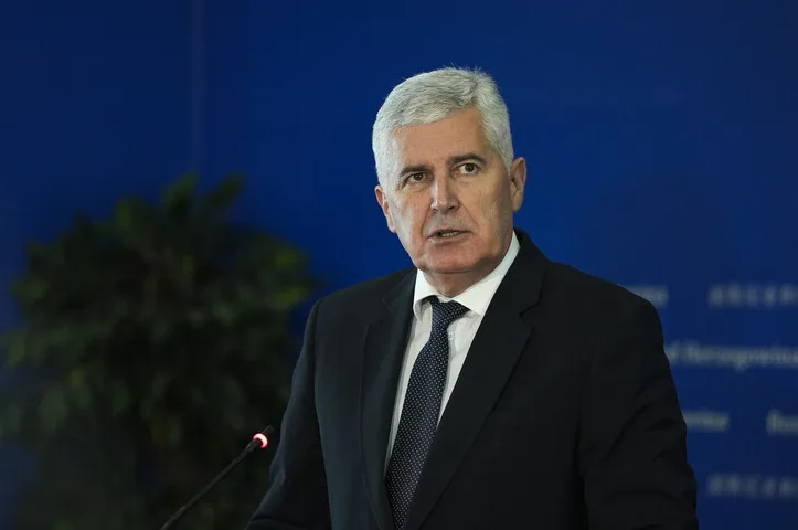 Političar sa sijedom kosom u crnom odijelu i plavoj kravati govori na konferenciji za medije ispred plave pozadine
