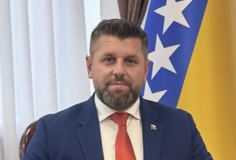 Portret muškarca u plavom odijelu i crvenoj kravati, sa bradom, ispred zastave Bosne i Hercegovine.