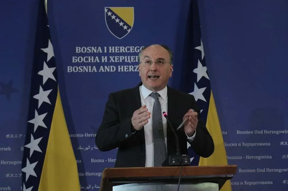 Političar iz Bosne i Hercegovine u odijelu i sa kravatom govori za govornicom, uz zastave Bosne i Hercegovine iza njega.