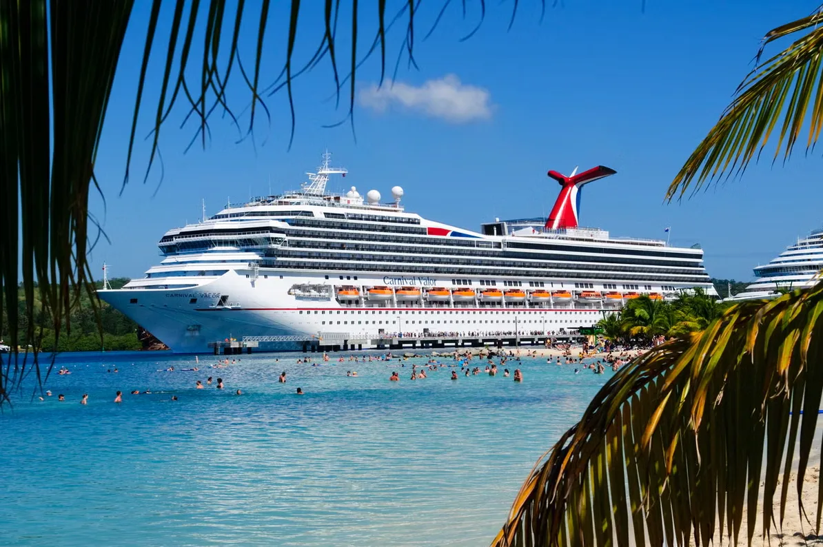 Veliki bijeli kruzer "Carnival Valor" usidren u plavom moru ispred tropske plaže sa mnogo ljudi koji plivaju i sunčaju se, sa palmama u prvom planu.
