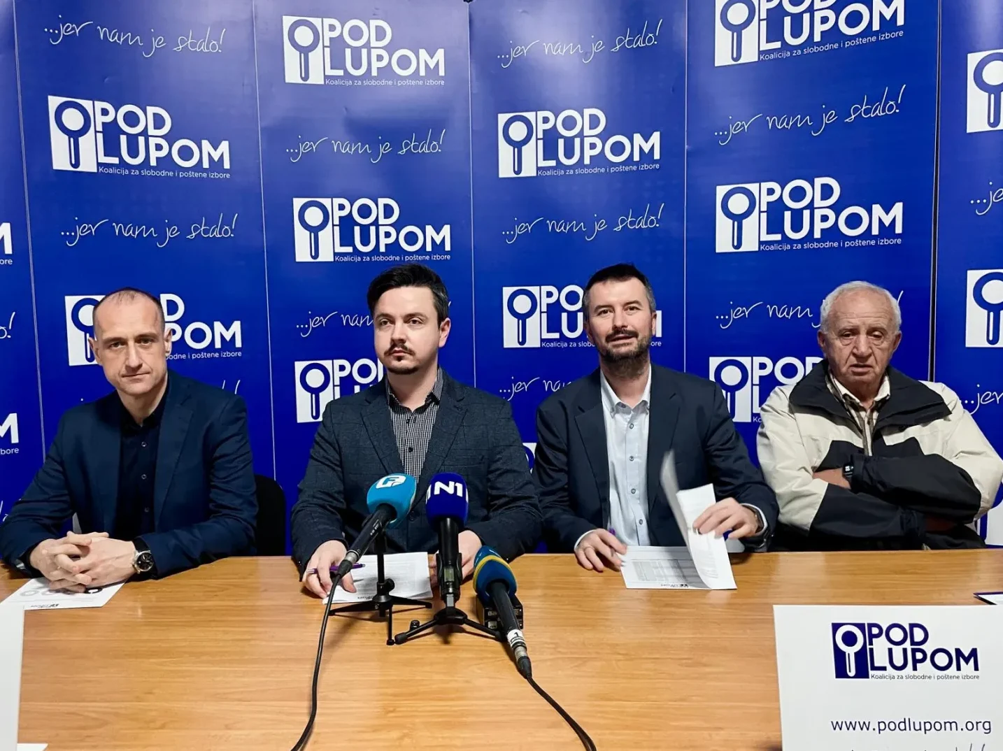 Četiri muškarca sjede za stolom ispred plave pozadine s logom "Pod Lupom".