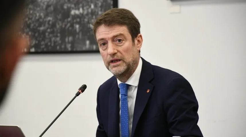 Portret muškarca u tamnom odijelu i plavoj kravati sa bijelim tačkicama, koji govori za govornicom.