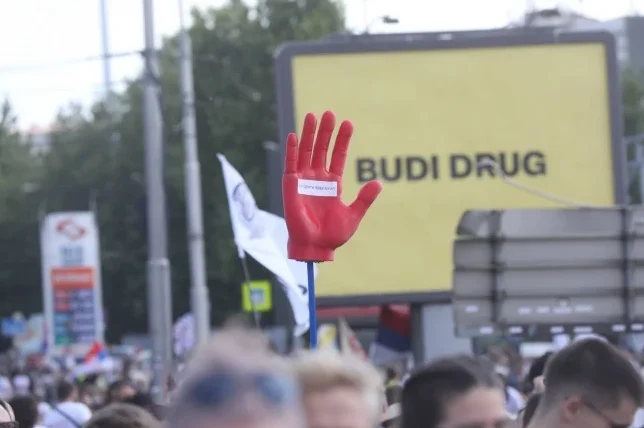 Crvena plastična ruka podignuta u zrak, na dlanu se nalazi natpis 'BUDI DRUG' ispisan na bijeloj podlozi, u pozadini se vidi žuti pano s natpisom 'BUDI DRUG' i mnoštvo ljudi na protestu.