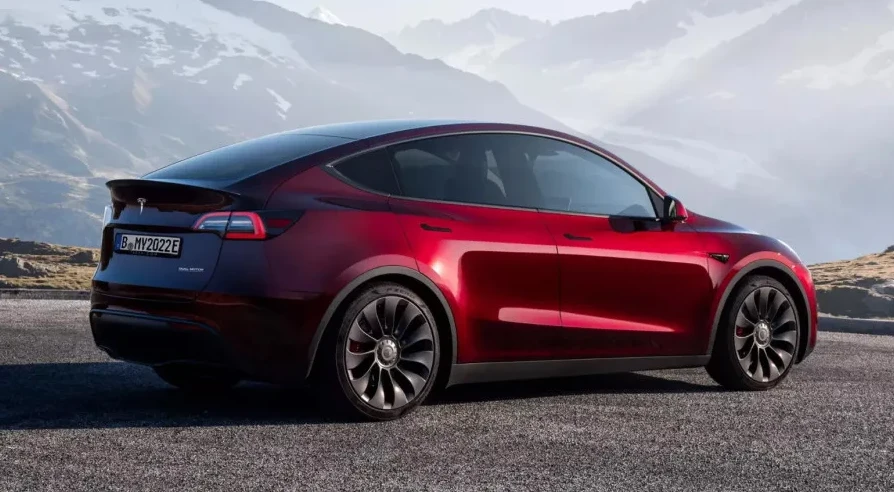 Crveni Tesla Model Y parkiran na asfaltu s planinskim lancem u pozadini.