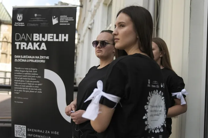 Tri djevojke nose crne majice sa bijelim trakama vezanim oko nadlaktica, stoje ispred panoa na kojem piše "DAN BIJELIH TRAKA - DAN SJEĆANJA NA ŽRTVE ZLOČINA U PRIJEDORU".