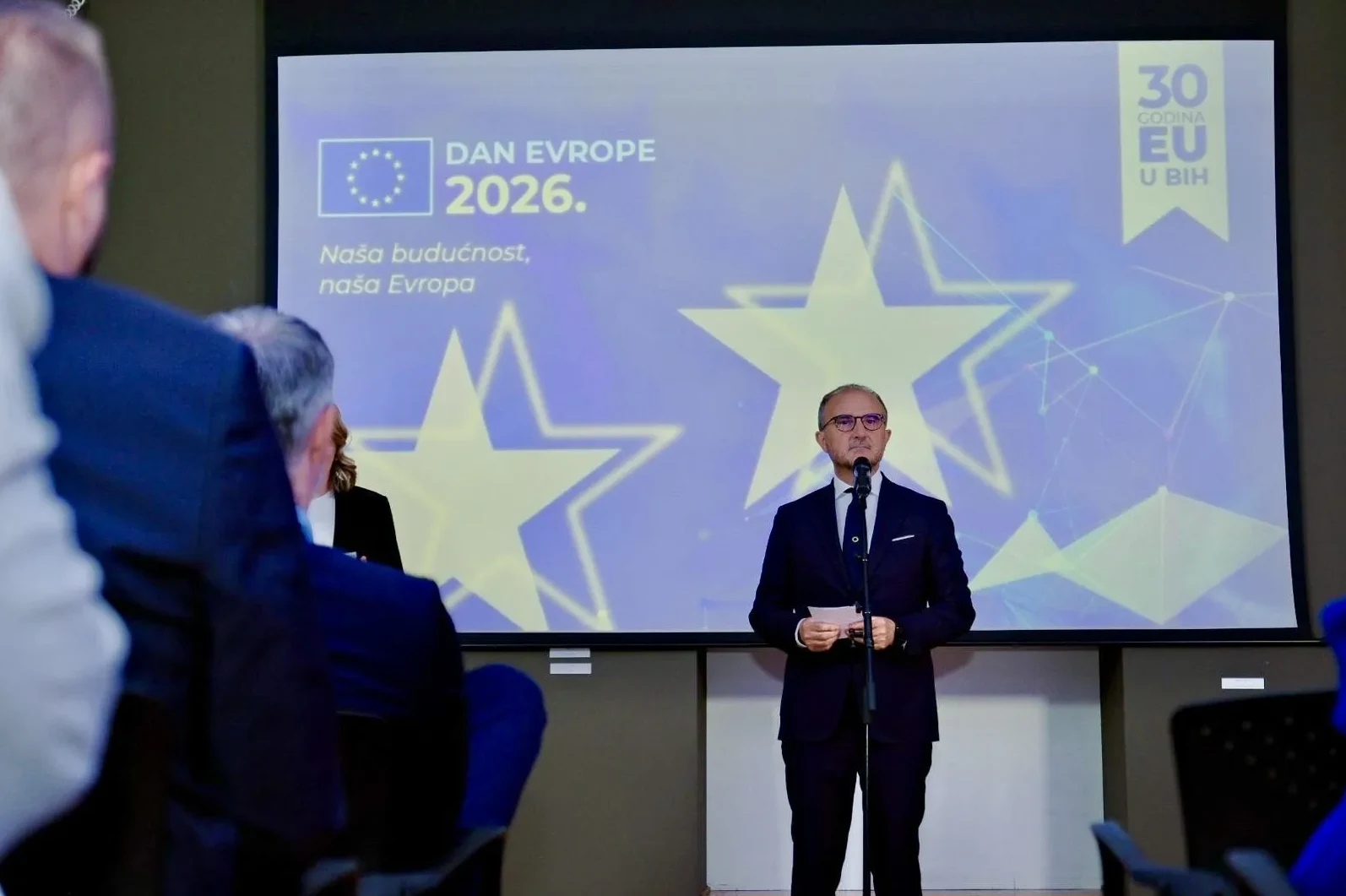 Političar drži govor pred publikom na pozornici, sa velikim ekranom u pozadini koji prikazuje logo "Dan Evrope 2026." i "30 Godina EU u BiH".