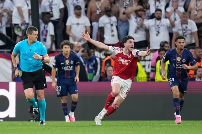 Fudbaler Arsenala Deklan Rajz (Declan Rice) u crvenom dresu sa brojem 40 postigao je gol, slaveći s podignutim rukama na zelenom travnjaku, dok ga u pozadini prate sudija i igrači Pari Sen Žermena (PSG) u tamnoplavim dresovima.