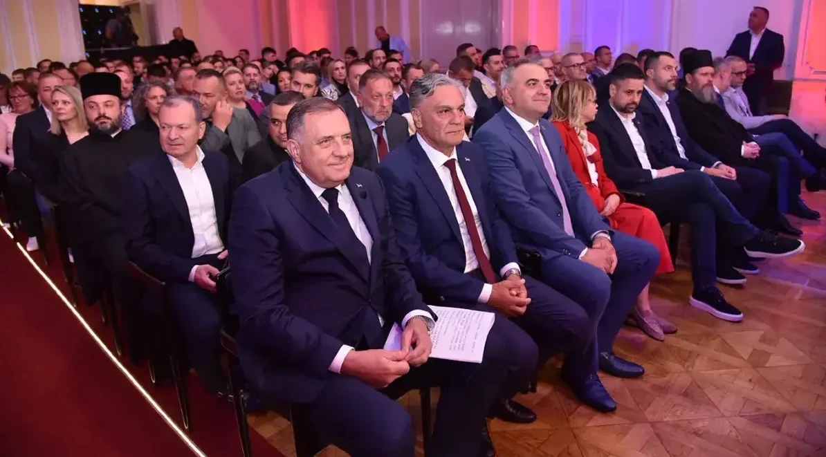Grupa muškaraca u odijelima sjedi u prvom redu na sjednici, dok ih gleda publika iza.