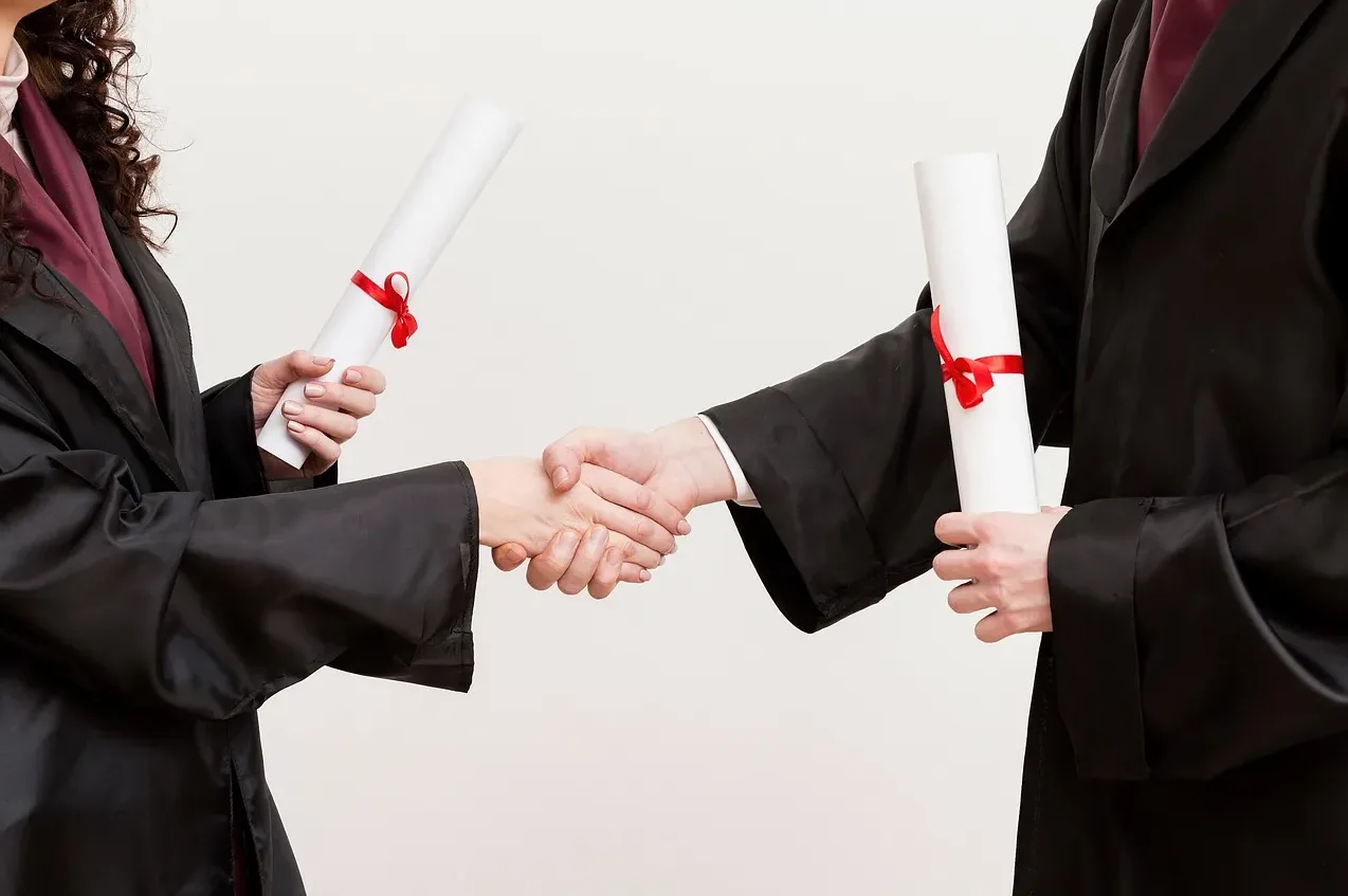 Dva diplomanta u akademskim odijelima rukuju se držeći diplome
