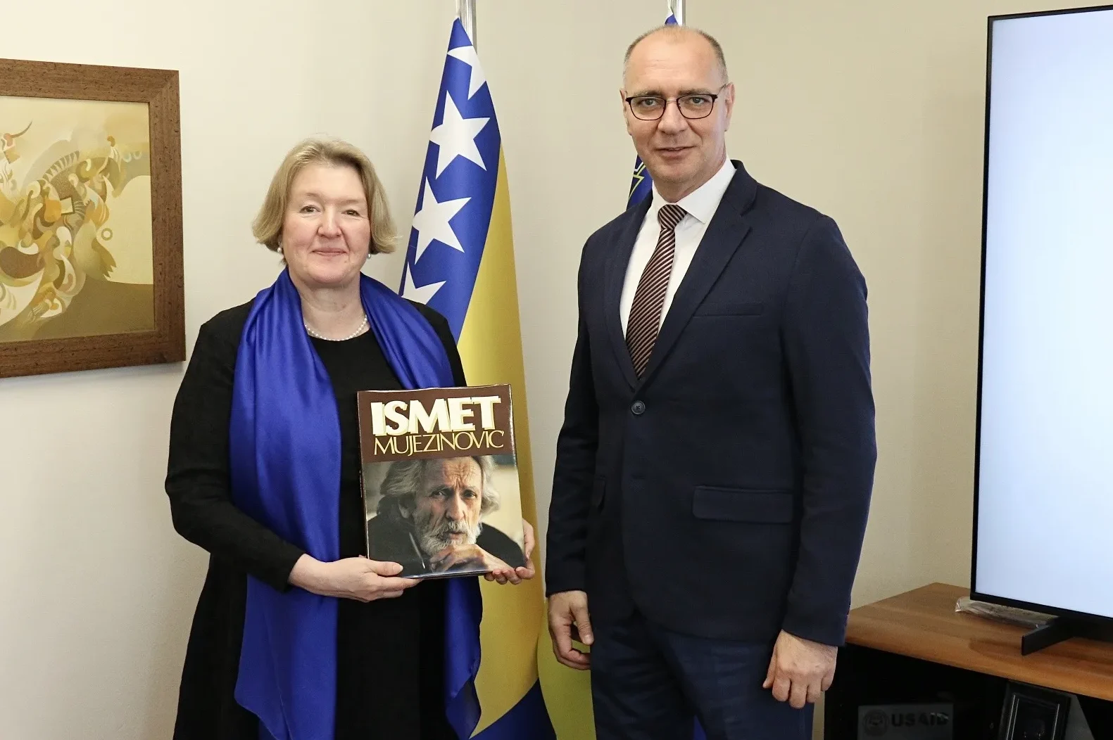 Žena i muškarac stoje ispred zastave Bosne i Hercegovine; žena drži knjigu sa portretom Ismeta Mujezinovića na naslovnici.