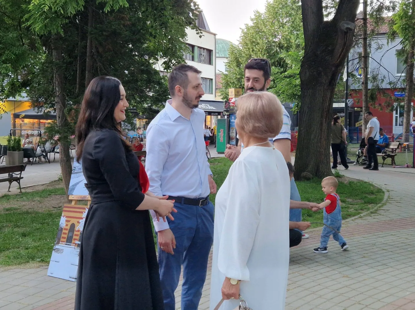 Grupa ljudi, uključujući djecu i odrasle, razgovara u parku pored drveća i zgrada.
