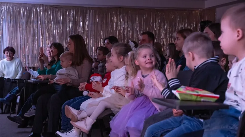 Djeca i odrasli sjede i gledaju nešto ispred sebe, neki od njih aplaudiraju.