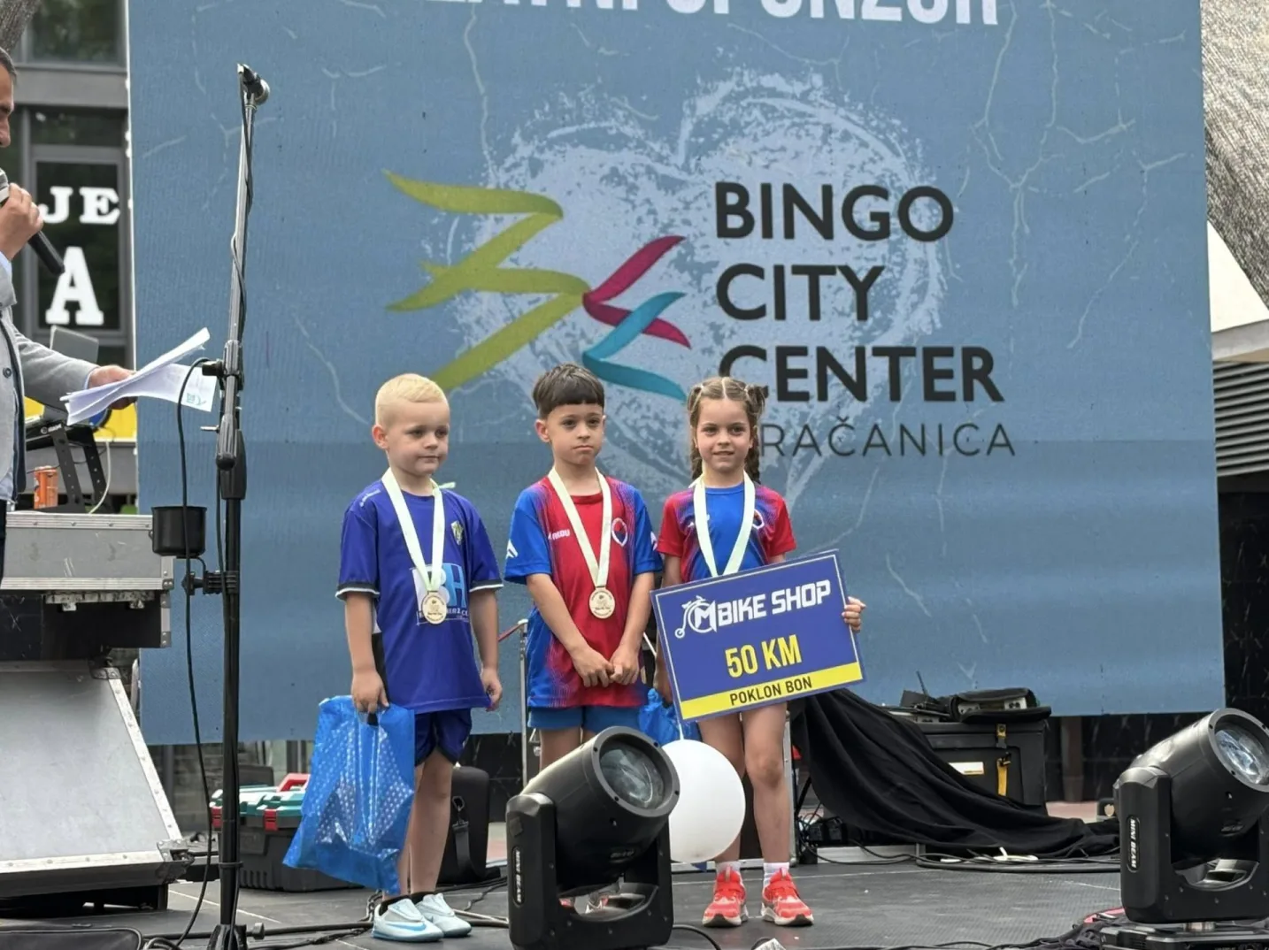 Tri djeteta, dva dječaka i jedna djevojčica, stoje na pozornici u sportskoj odjeći i sa medaljama oko vrata. Djevojčica drži znak "Bike Shop 50 KM Poklon Bon". U pozadini je pano sa logom "Bingo City Center" i "Gradačac".