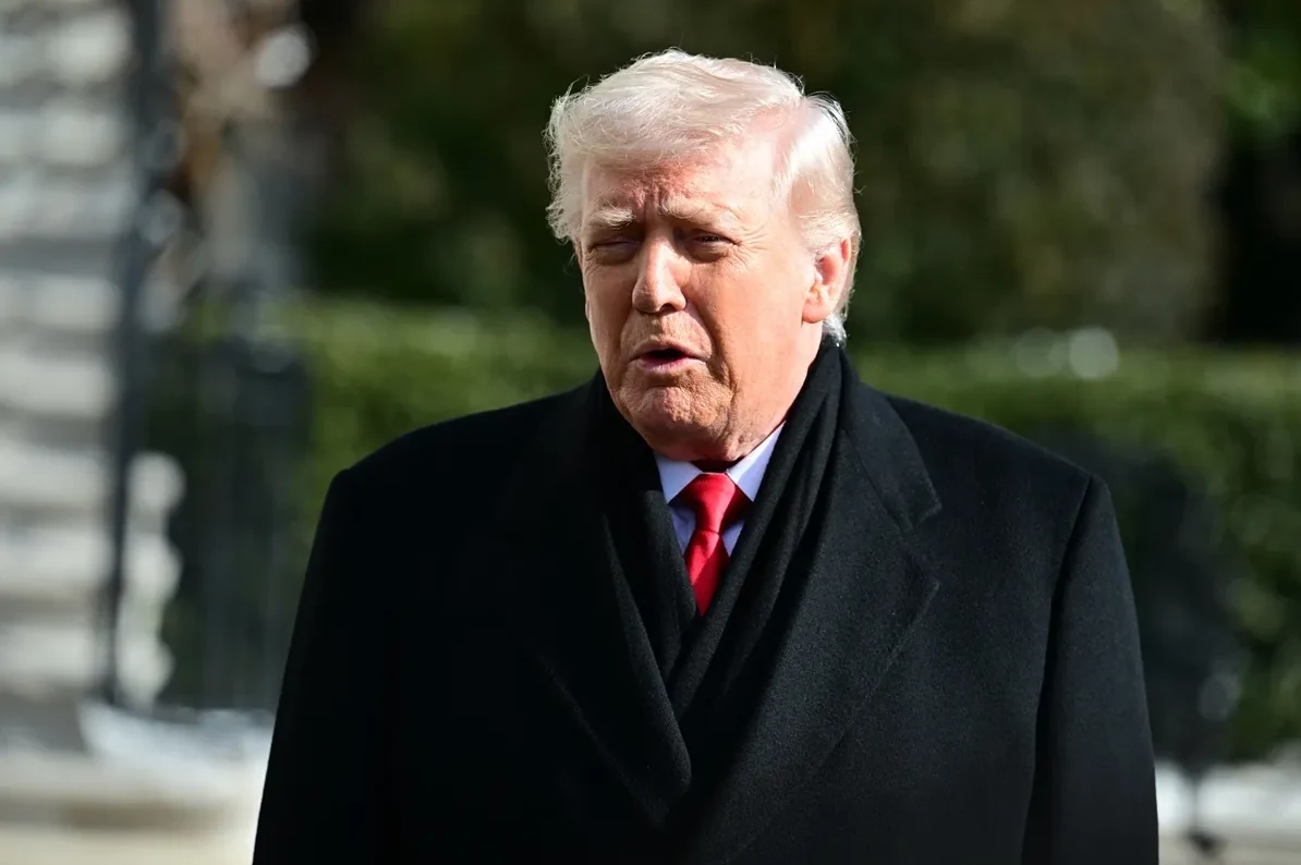 Donald Trump u crnom kaputu i crvenoj kravati, sa bijelom kosom i zatvorenim očima
