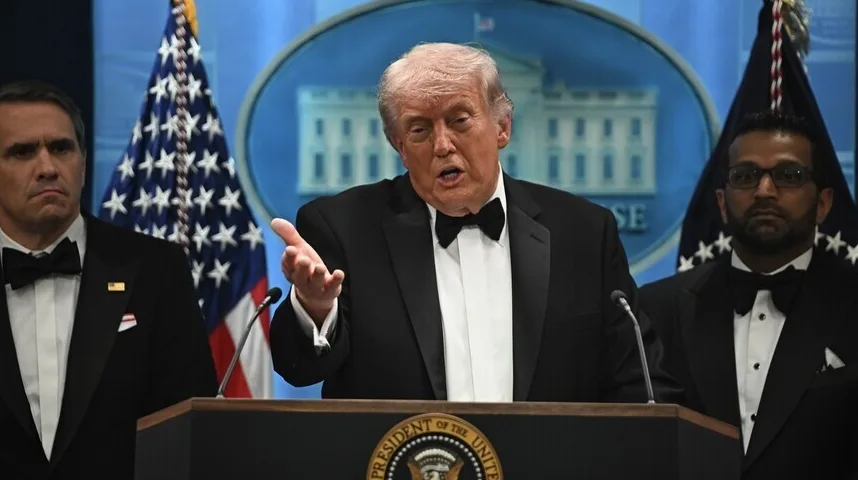 Donald Trump u crnom smokingu s leptir-mašnom govori za govornicom, pored njega stoje dva muškarca u sličnoj odjeći.