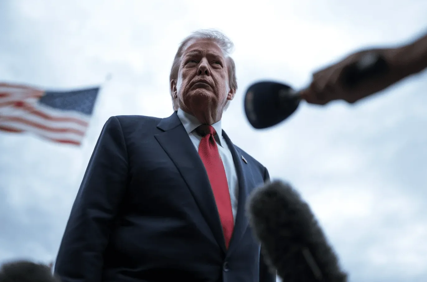 Donald Trump u tamnom odijelu i crvenoj kravati, sa američkom zastavom u pozadini, dok mu novinari postavljaju pitanja.