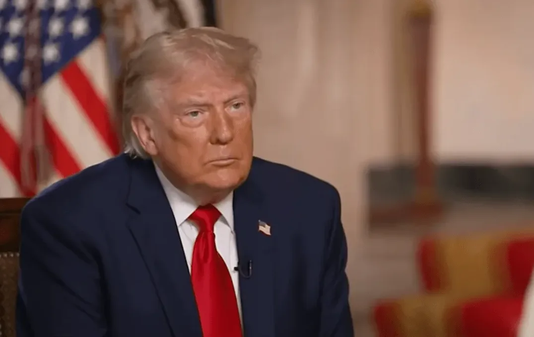 Donald Trump u tamnoplavom odijelu i crvenoj kravati, sjedi pred američkom zastavom.