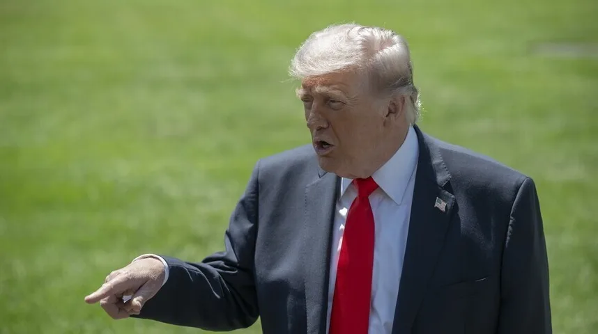Donald Trump, bivši predsjednik Sjedinjenih Američkih Država, u tamnom odijelu i crvenoj kravati, sa podignutim kažiprstom u gestikulaciji, stoji na zelenom travnjaku.
