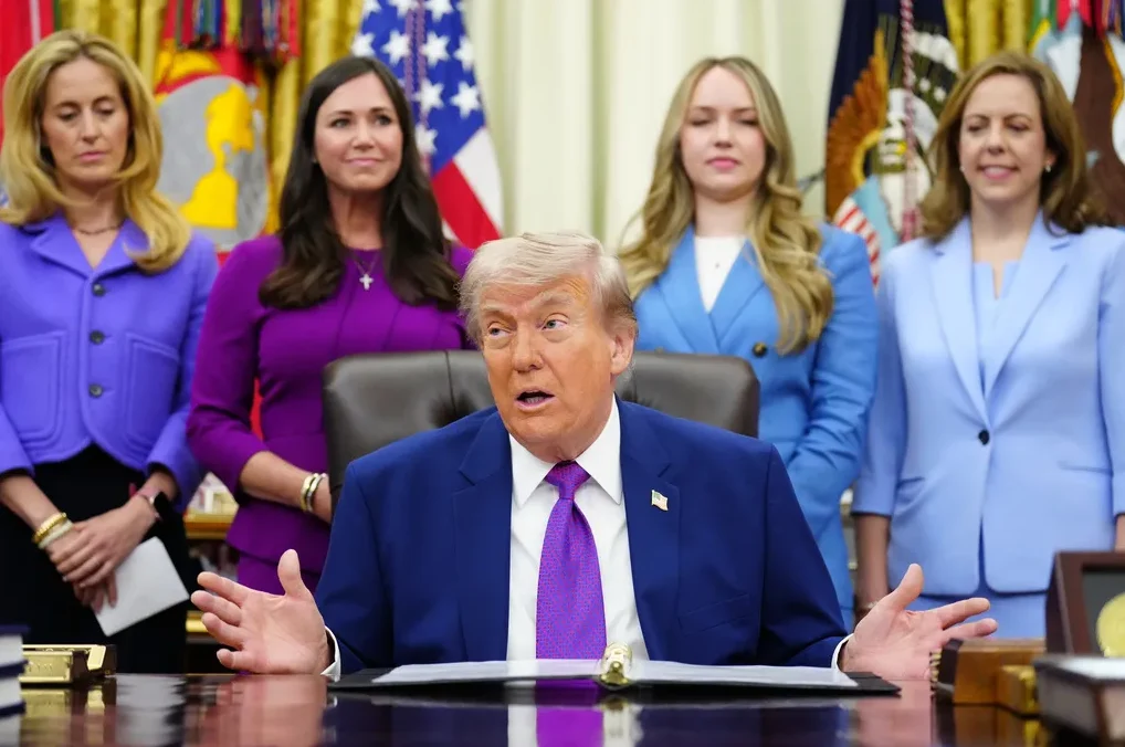Donald Trump sjedi za stolom u Ovalnom uredu, govoreći i gestikulirajući rukama, dok iza njega stoje četiri žene odjevene u plave i ljubičaste kostime.