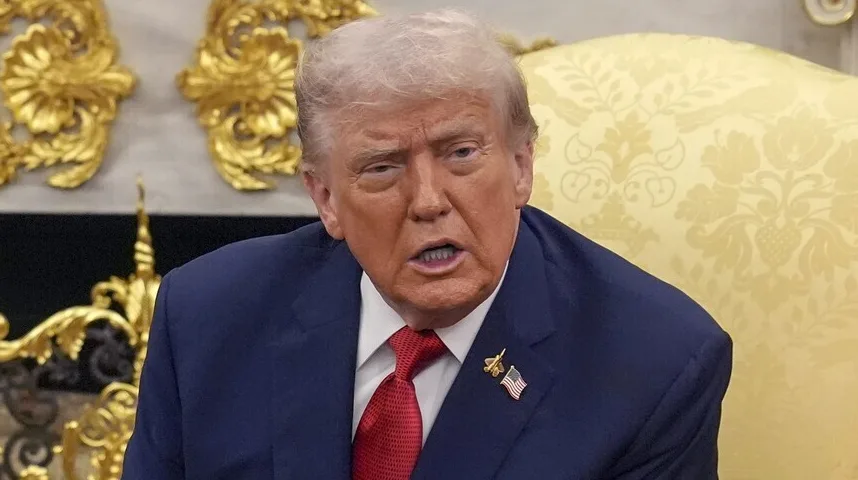 Donald Trump u tamnoplavom odijelu i crvenoj kravati, s američkom zastavicom na reveru, govori u zatvorenom prostoru s pozadinom ukrašenom zlatnim ornamentima.