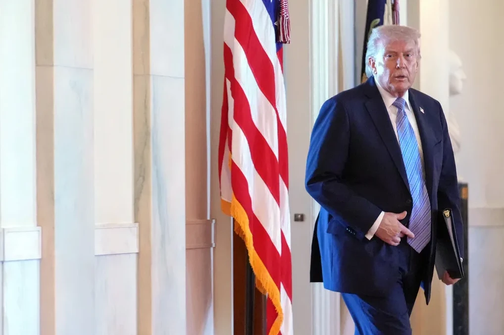 Donald Trump u tamnom odijelu s plavom prugastom kravatom hodajući pored američke zastave.