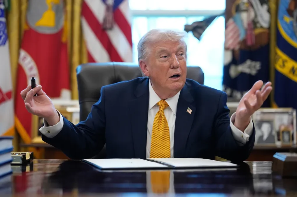 Donald Trump sjedi za radnim stolom u Ovalnom uredu, drži olovku i gestikulira.