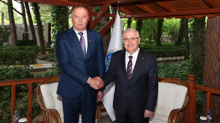 Dva ugledna gospodina u odijelima rukuju se ispred zastave u vanjskom prostoru okruženom zelenilom.