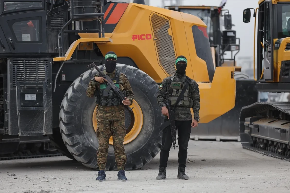 Dva naoružana muškarca u vojnim uniformama i maskiranim licima stoje ispred velikih žutih građevinskih mašina.
