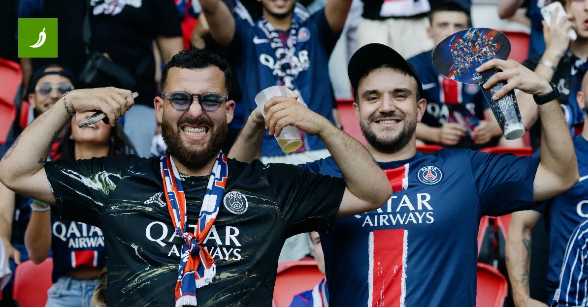 Dva nasmijana muška navijača Paris Saint-Germaina na stadionu, jedan drži piće, a drugi telefon, obojica nose šalove.