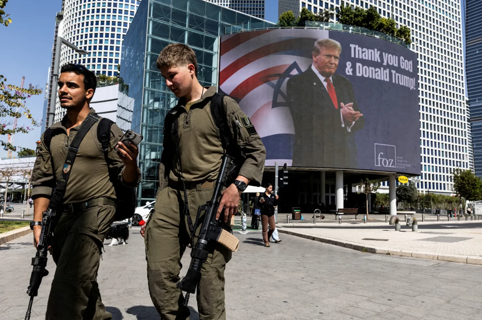 Dva naoružana vojnika u maslinastozelenim uniformama hodaju ulicom ispred velikog digitalnog ekrana na kojem se vidi lik Donalda Trumpa sa porukom 'Thank you God & Donald Trump!' i zastavom Izraela.