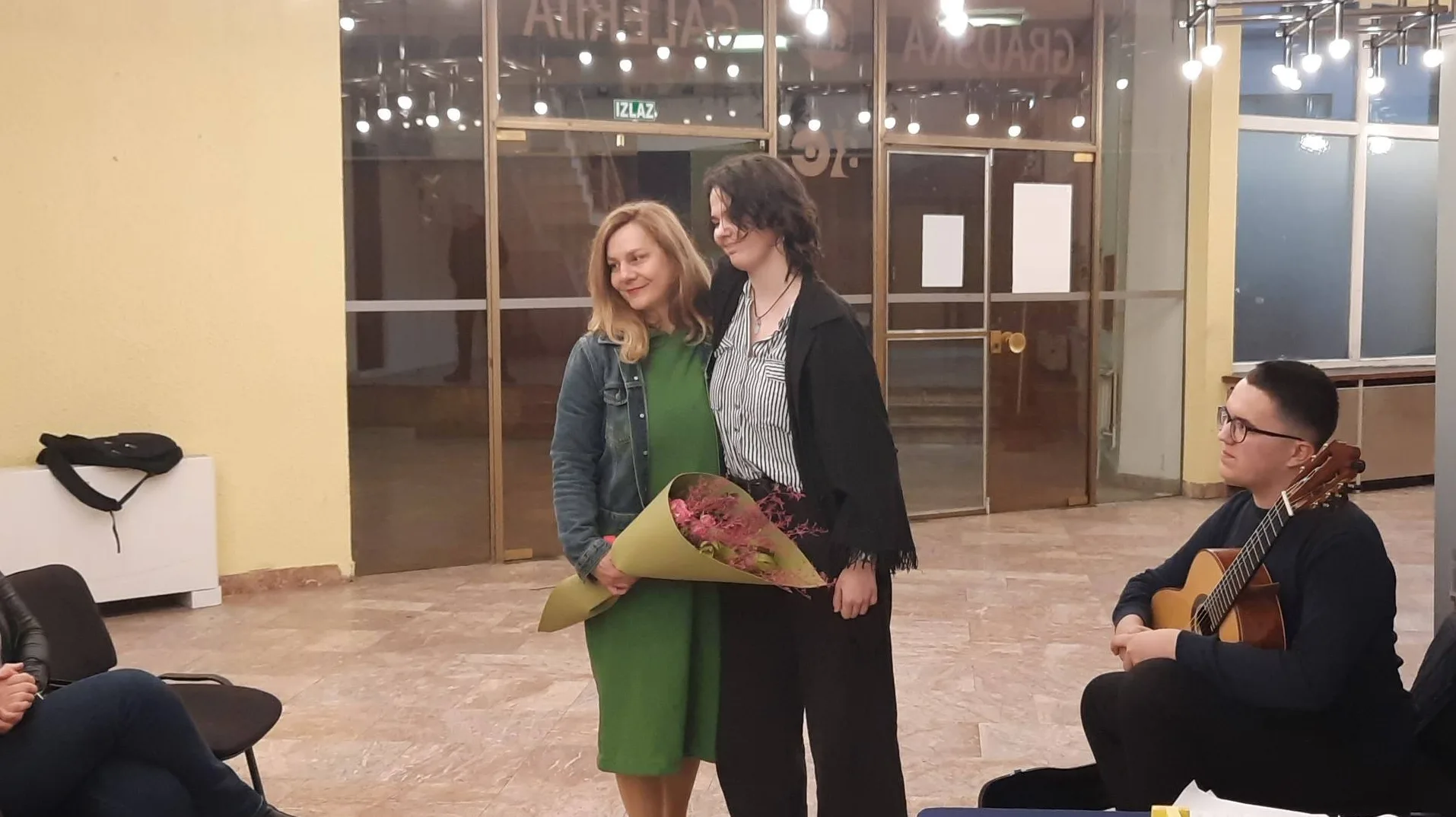 Dvije žene, jedna drži buket cvijeća, nasmijane stoje zajedno. U pozadini se vidi natpis "IZLAZ", a sa desne strane sjedi mladić s gitarom.