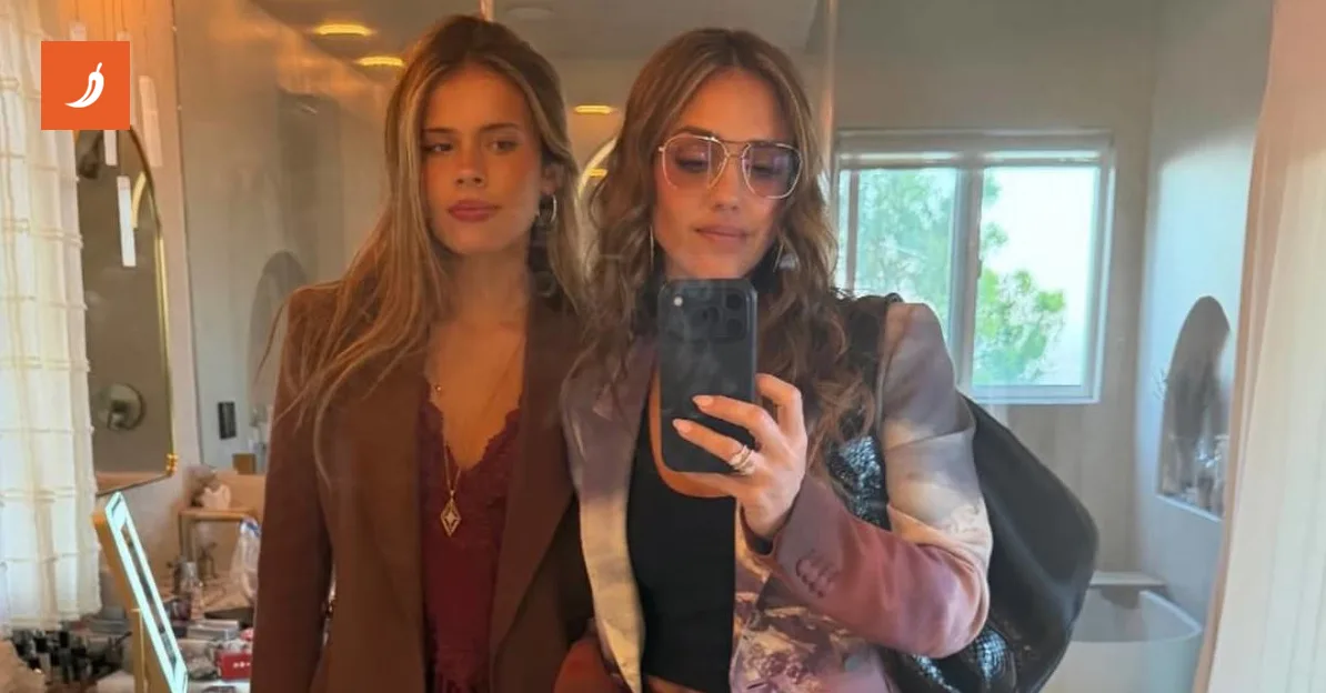 Dvije žene, majka i kći, poziraju za selfie u kupaonici. Starija žena, vjerovatno majka, nosi naočale i šareni sako, dok mlađa žena nosi smeđi sako.