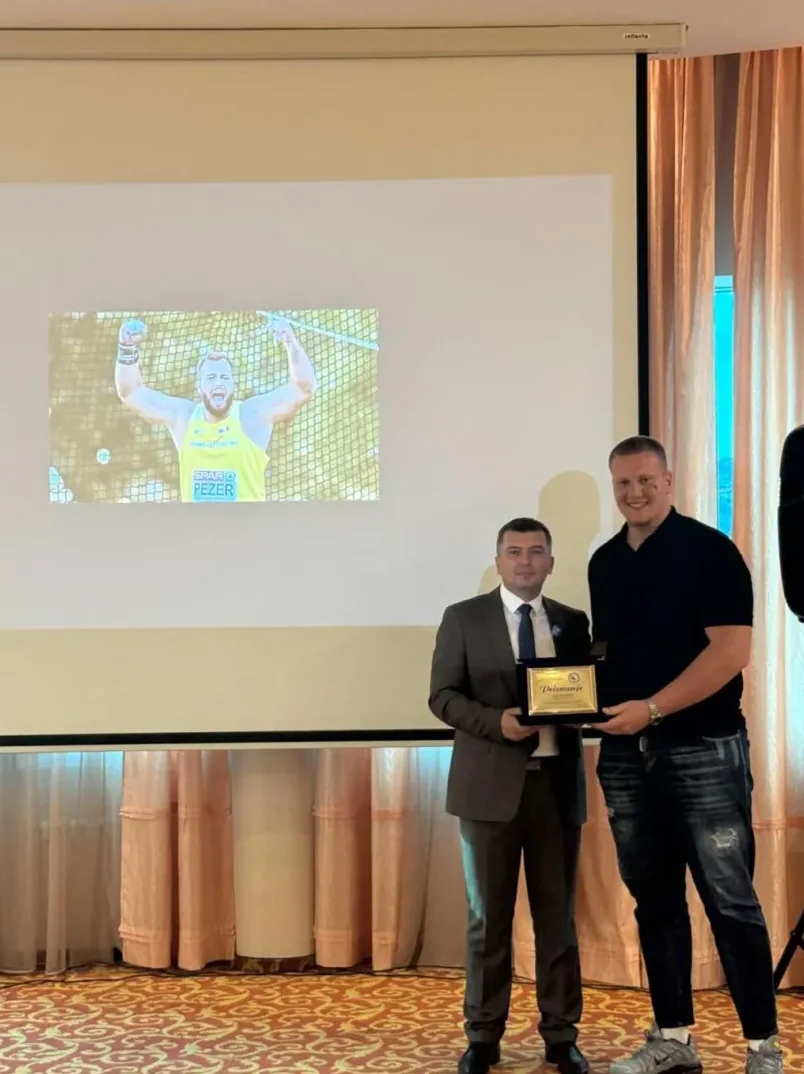 Dva muškarca stoje ispred projekcionog platna na kojem je prikazan atletičar koji drži teg i slavi pobjedu. Jedan od muškaraca, obučen u odijelo, uručuje zlatnu plaketu drugom muškarcu obučenom u crnu majicu.