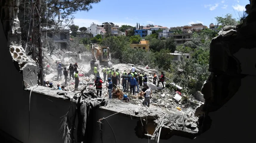 Grupa spasilaca i radnika u žutim prslucima okružuje iskopani rov pored uništene zgrade, dok bager vrši iskopavanja na terenu prekrivenom šutom.