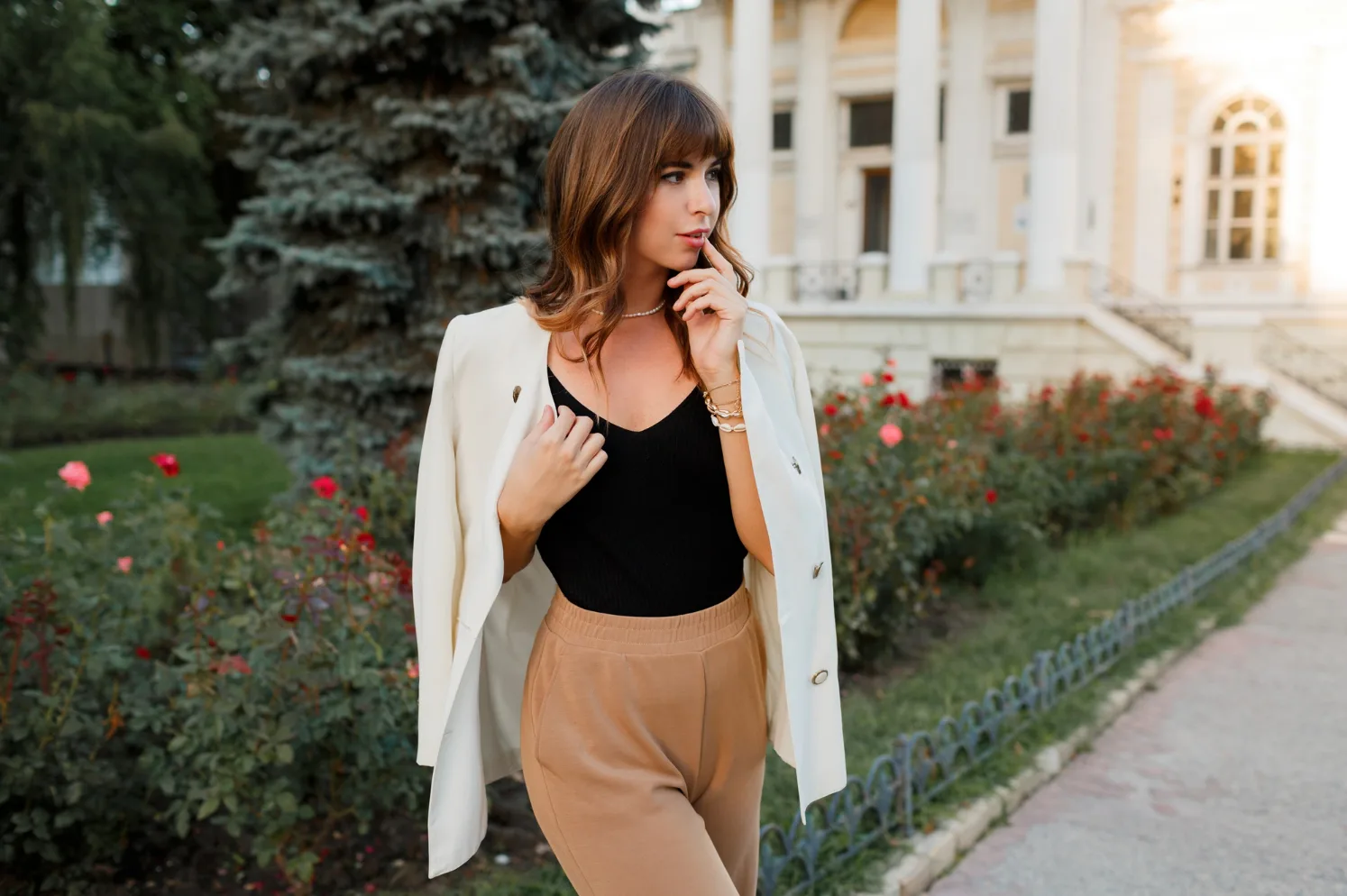 Elegantna mlada žena u crnom topu, bijelom sakou i braon pantalonama, sa bisernom ogrlicom i narukvicama, pozira u parku ispred zgrade sa stubovima, okružena zelenilom i cvijećem.