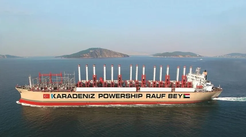 Brod za proizvodnju električne energije, nazvan Karadeniz Powership Rauf Bey, plovi kroz plavo more ispred obale sa zelenim brdima u pozadini.