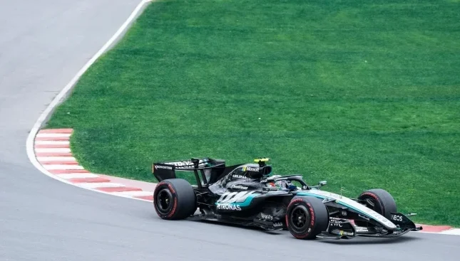 Crni bolid Formule 1 sa zelenim detaljima, sa logom "PETRONAS" istaknutim sa strane, kako prolazi kroz blagu krivinu na asfaltnoj stazi pored zelene trave.