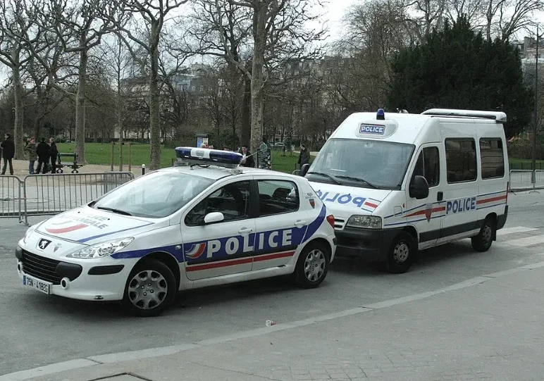 Dva bijela policijska vozila, automobil i kombi, parkirana su uz cestu u Parizu. Automobil marke Peugeot ima plavo-crvenu traku s natpisom "POLICE" i rotacijsko svjetlo na krovu. Kombi, koji je iza automobila, također ima natpis "POLICE" i plavo svjetlo. U pozadini se vide stabla bez lišća i zgrada.