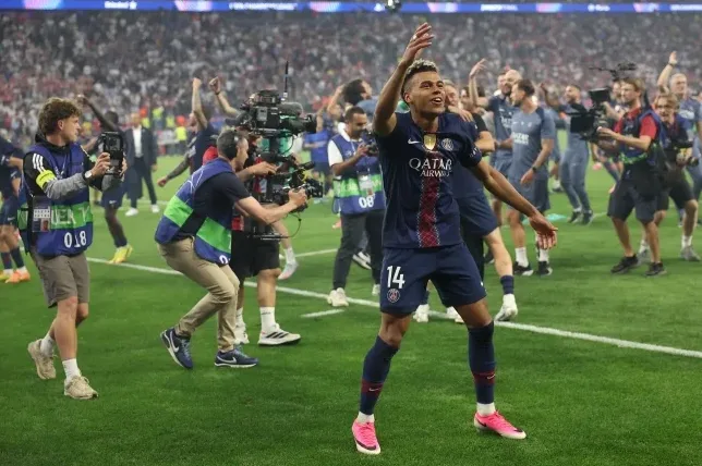 Fudbaler PSG-a, sa brojem 14 na dresu, slavi na terenu uz podignutu ruku, okružen medijskim radnicima i saigračima u pozadini.