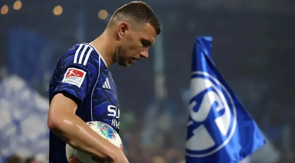 Fudbaler u plavom dresu sa amblemom Bundeslige drži loptu, dok se u pozadini vidi plava zastava sa bijelim grbom kluba.