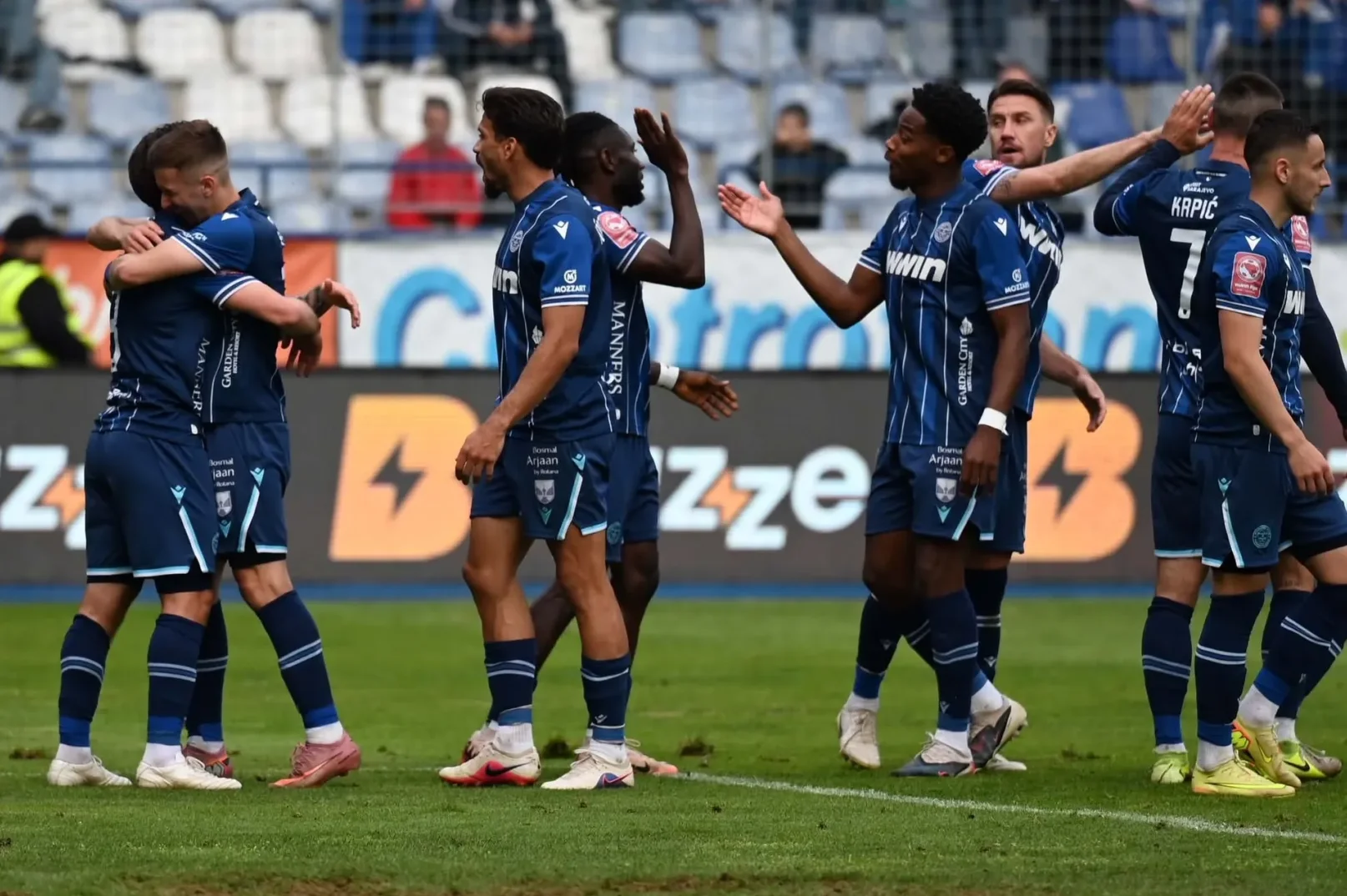 Fudbaleri u plavim dresovima grle se i čestitaju jedni drugima na terenu, slaveći postignut gol.