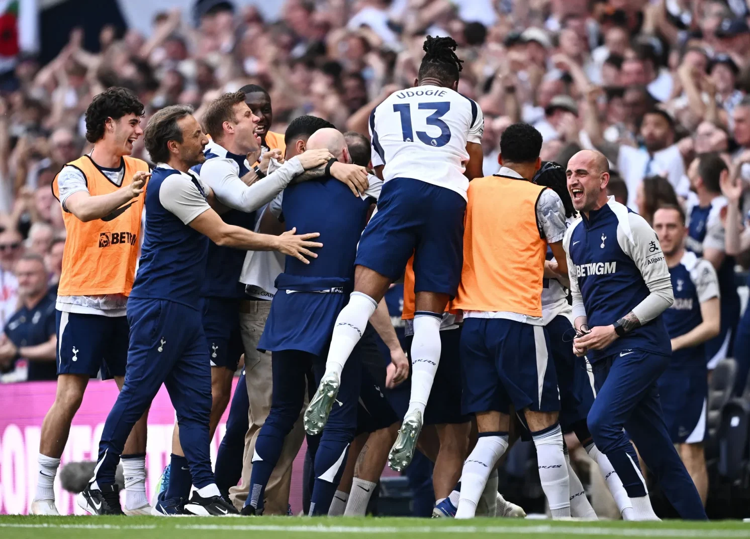 Fudbaleri Tottenhama slave pogodak na terenu, sa trenerima i saigračima okruženi navijačima u pozadini.