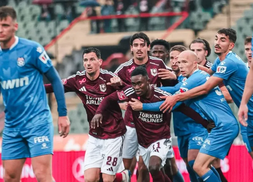 Fudbaleri u tamno-crvenim i plavim dresovima vode borbu za loptu na fudbalskom stadionu.