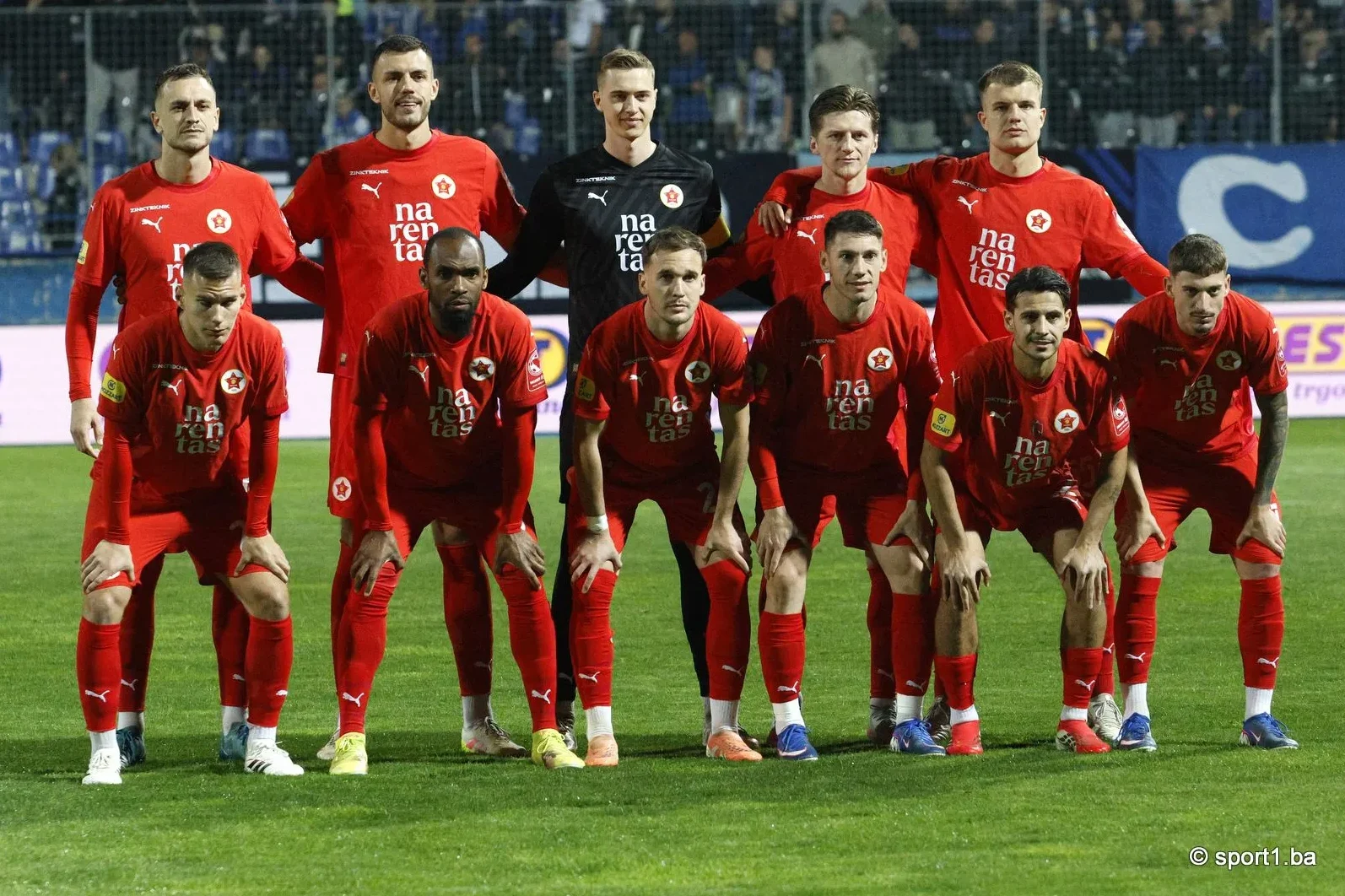 Fudbalska ekipa u crvenim dresovima pozira na zelenom terenu.