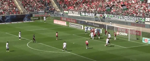 Fudbalski igrači u bijelim i crnim dresovima bore se za loptu u kaznenom prostoru pred golom.