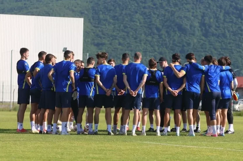 Fudbalski tim okupljen u krugu na zelenom terenu, obučeni u plave dresove i tamne šorceve.