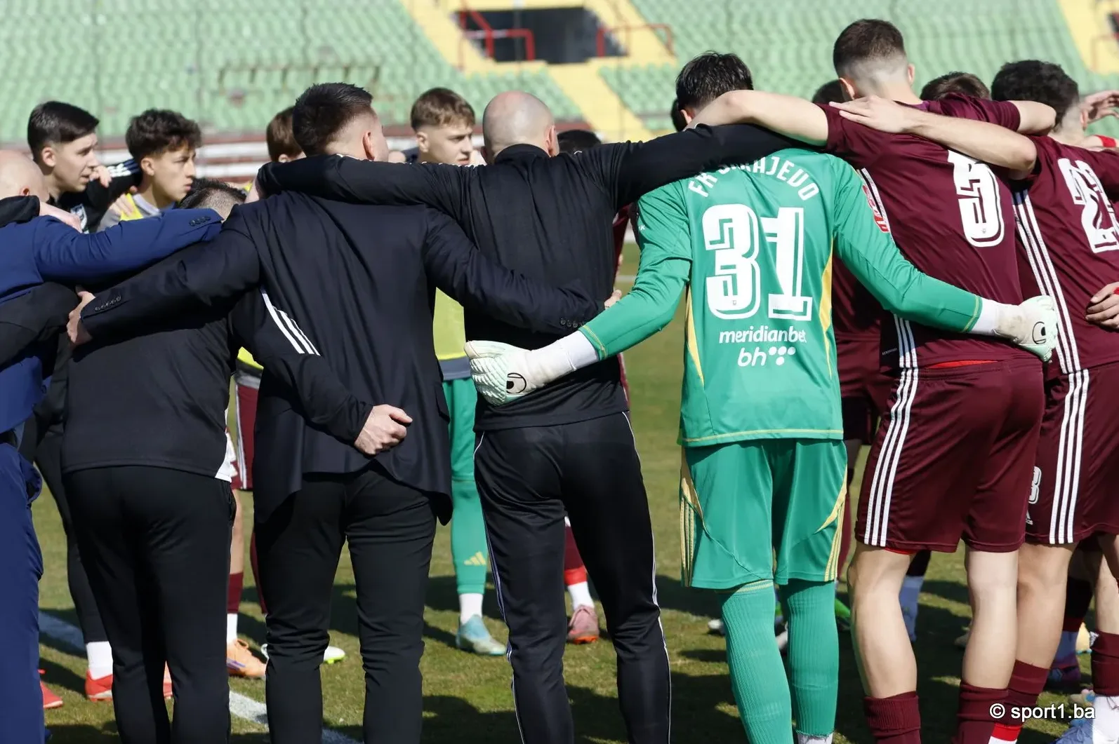 Fudbalski tim okupljen u zagrljaju na terenu, sa fokusom na igrače i golmana.