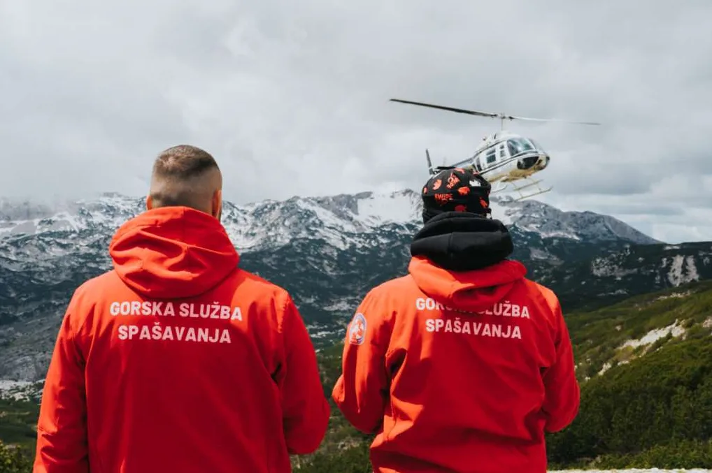 Dva člana Gorske službe spasavanja, odjevena u crvene jakne sa natpisom "GORSKA SLUŽBA SPAŠAVANJA", stoje okrenuta leđima posmatrajući helikopter koji leti iznad planinskog predjela.