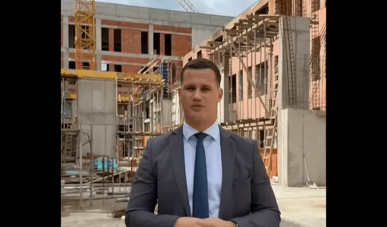 Mladić u odijelu i plavoj kravati stoji ispred zgrade u izgradnji sa dizalicom u pozadini.